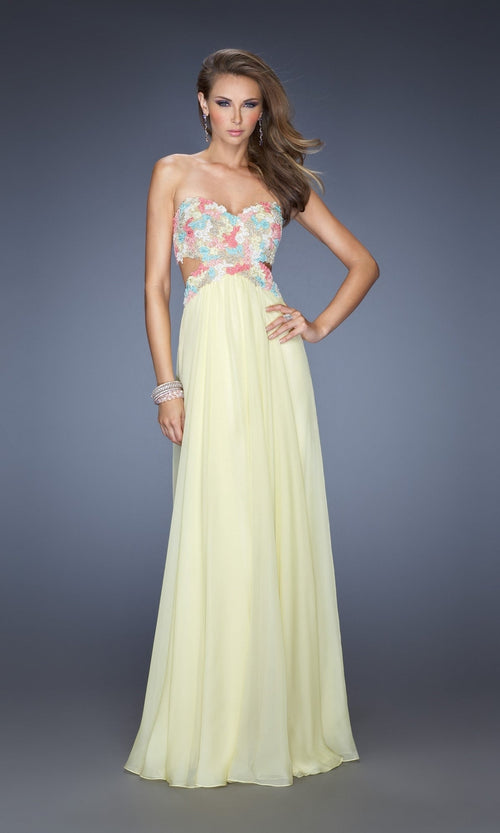 A woman models the La Femme Multicolor-Lace Chiffon Prom Dress 20059, a strapless, floor-length yellow gown with floral lace bodice and sheer side cutouts, standing before a plain gray background.