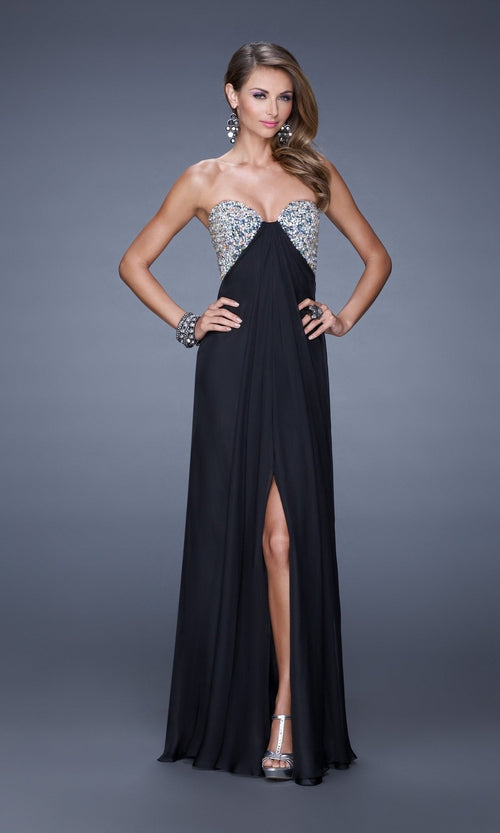 A woman stands wearing the La Femme Dress 20784, a strapless black chiffon gown with an embellished bust and front slit, accessorized with earrings, bracelets, and silver heels.