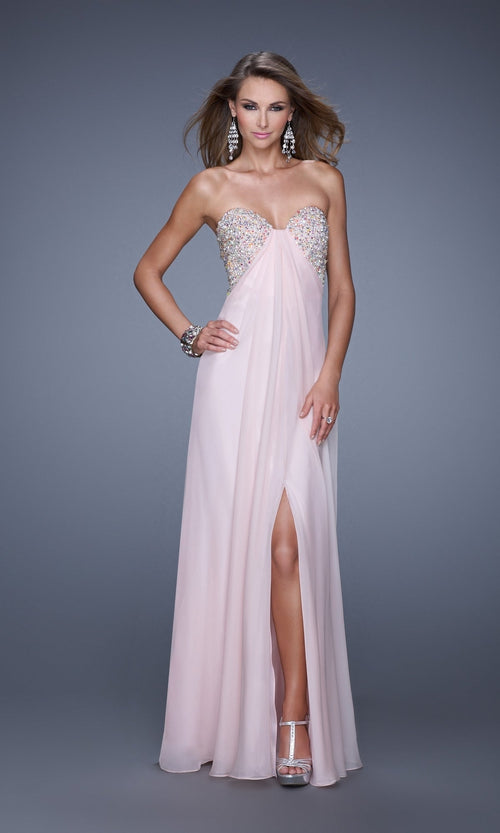 A woman in the La Femme Dress 20784, a strapless floor-length light pink chiffon gown with an embellished bust and front slit, pairs it with silver heels, statement earrings, and a bracelet against a gray background.
