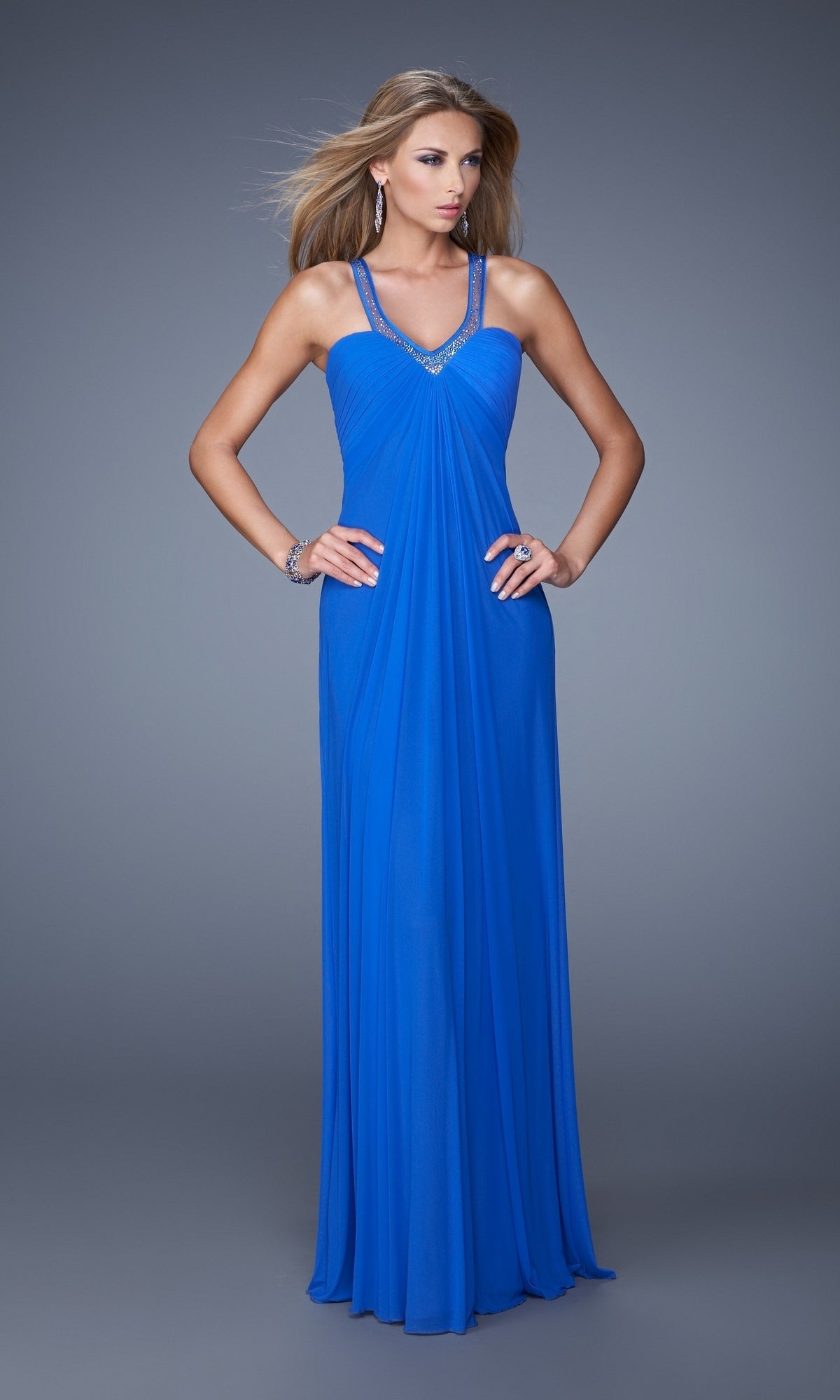 A woman poses against a gray background, hands on hips, wearing the La Femme Dress 20903â€”a floor-length royal blue gown with embellished straps.