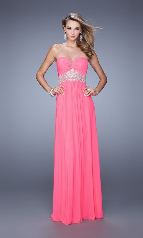 A woman wearing the Strapless La Femme Ruched Designer Prom Dress 21357â€”a floor-length pink gown with silver waist embellishments and subtle ruchingâ€”stands against a plain gray background.