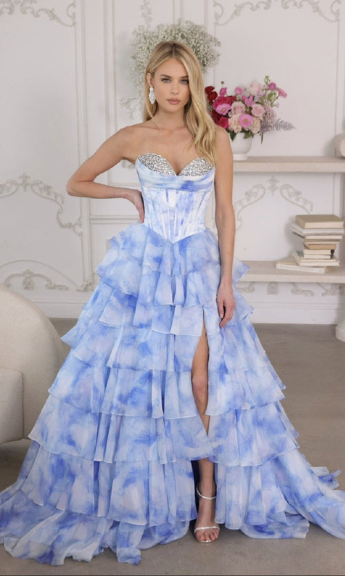 A woman stands indoors by shelves with books and flowers, wearing the Ava Presley 26442 long prom dress—strapless blue and white corset bodice, tiered ruffle skirt, and high slit.