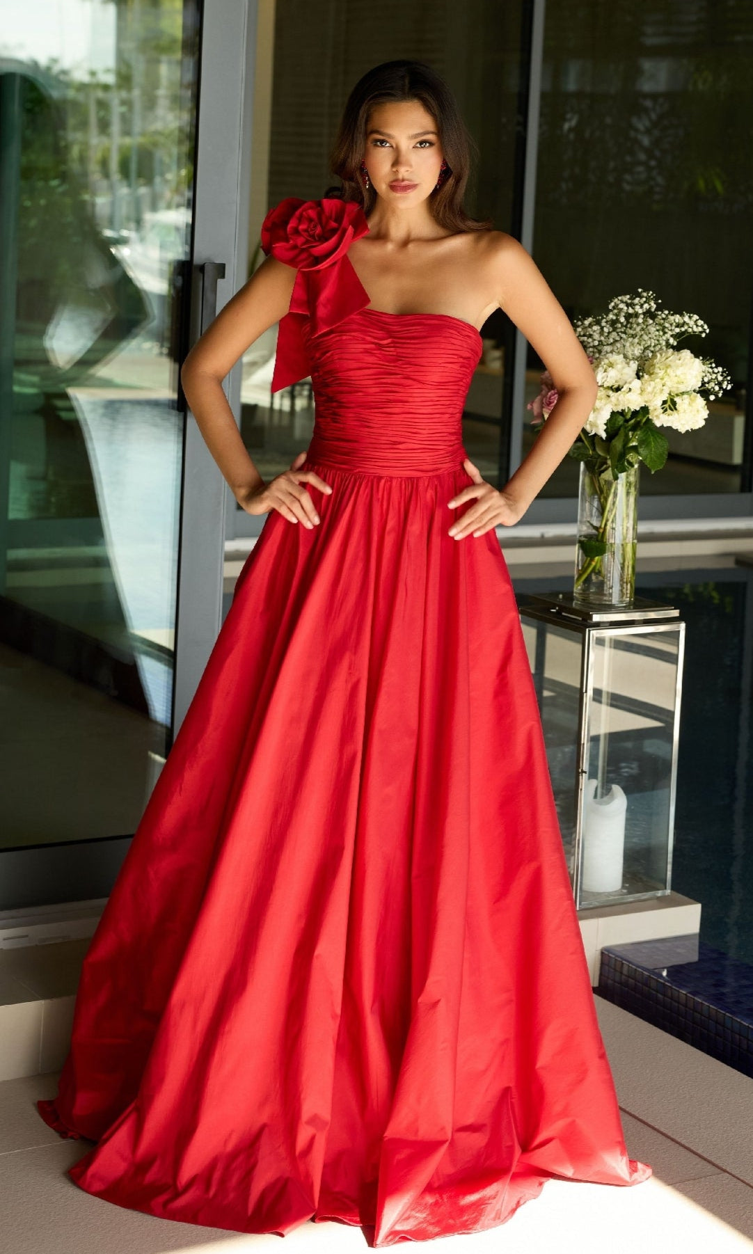 A woman in the Ava Presley 26524 long prom dress, a red one-shoulder gown with a floral accent on the shoulder, stands indoors by a glass door and a vase of flowers.