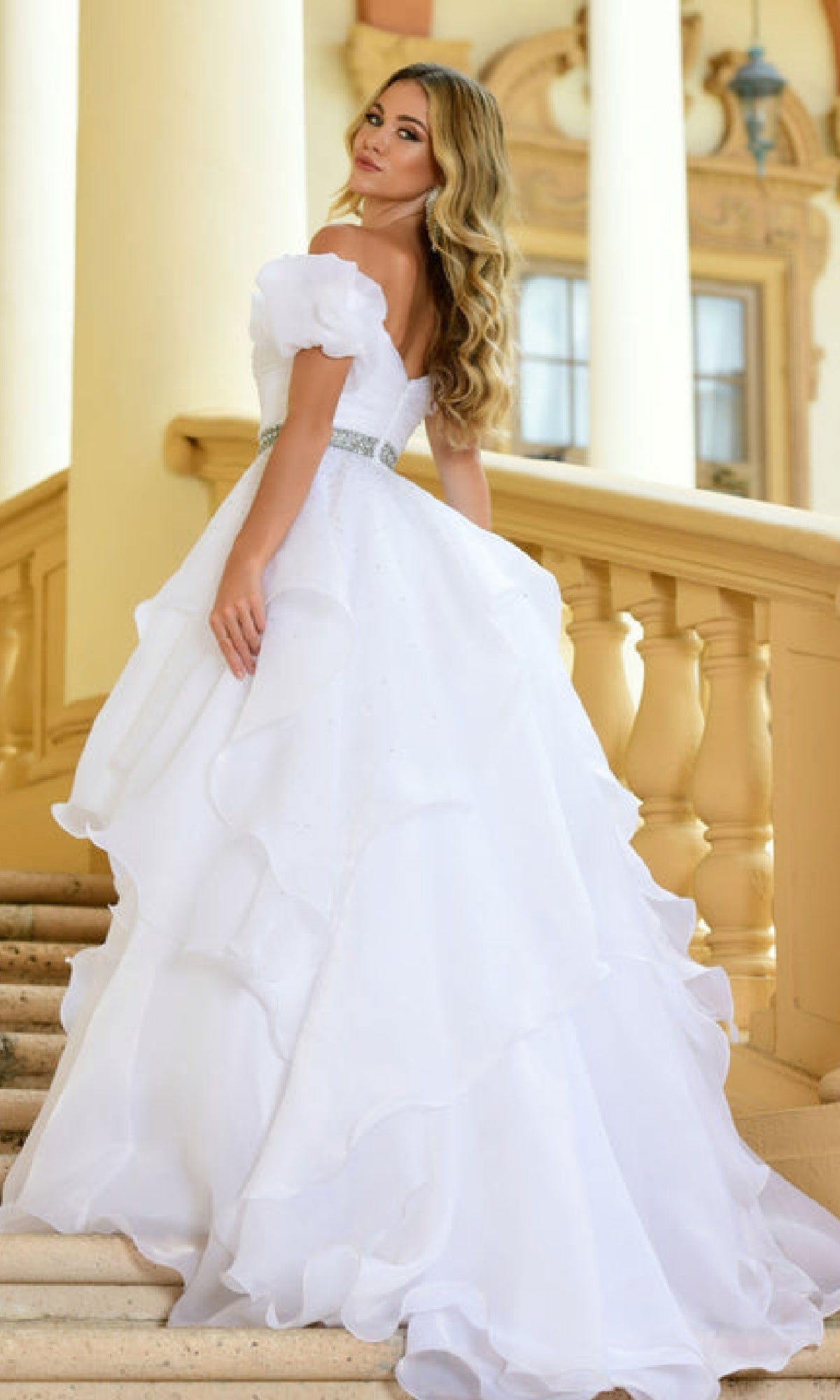 A woman in the Ava Presley 28571 puff-sleeve long prom ball gown stands on a staircase, looking over her shoulder.