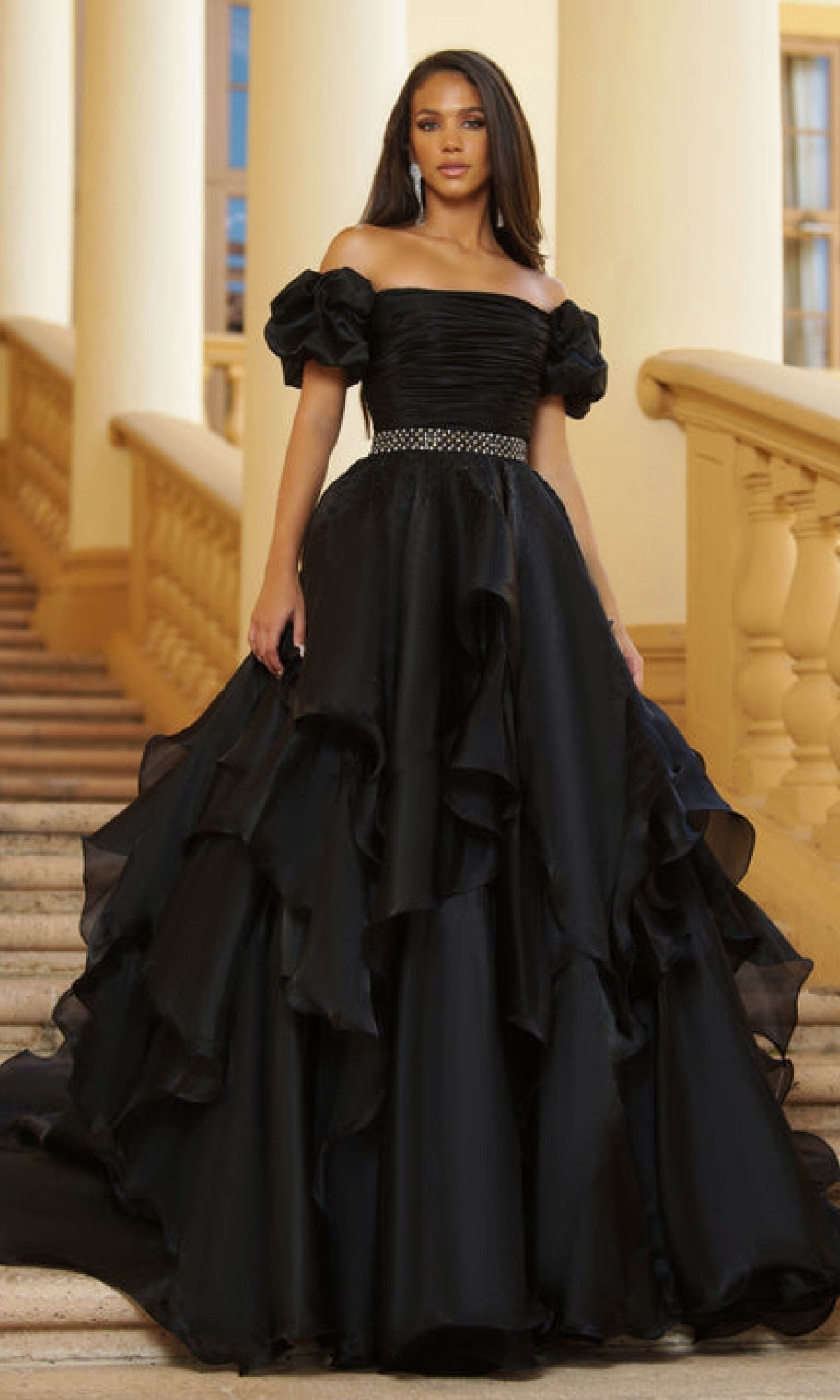 A woman stands on stairs in front of a building, wearing the Ava Presley 28571 Puff-Sleeve Long Prom Ball Gown with off-the-shoulder ruffled black layers and a jeweled belt.