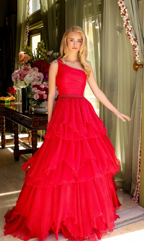 A woman stands indoors wearing the Ava Presley 29131 One-Shoulder Tiered Prom Gown, a vibrant red dress with beaded accents, framed by green curtains and a floral-adorned table.
