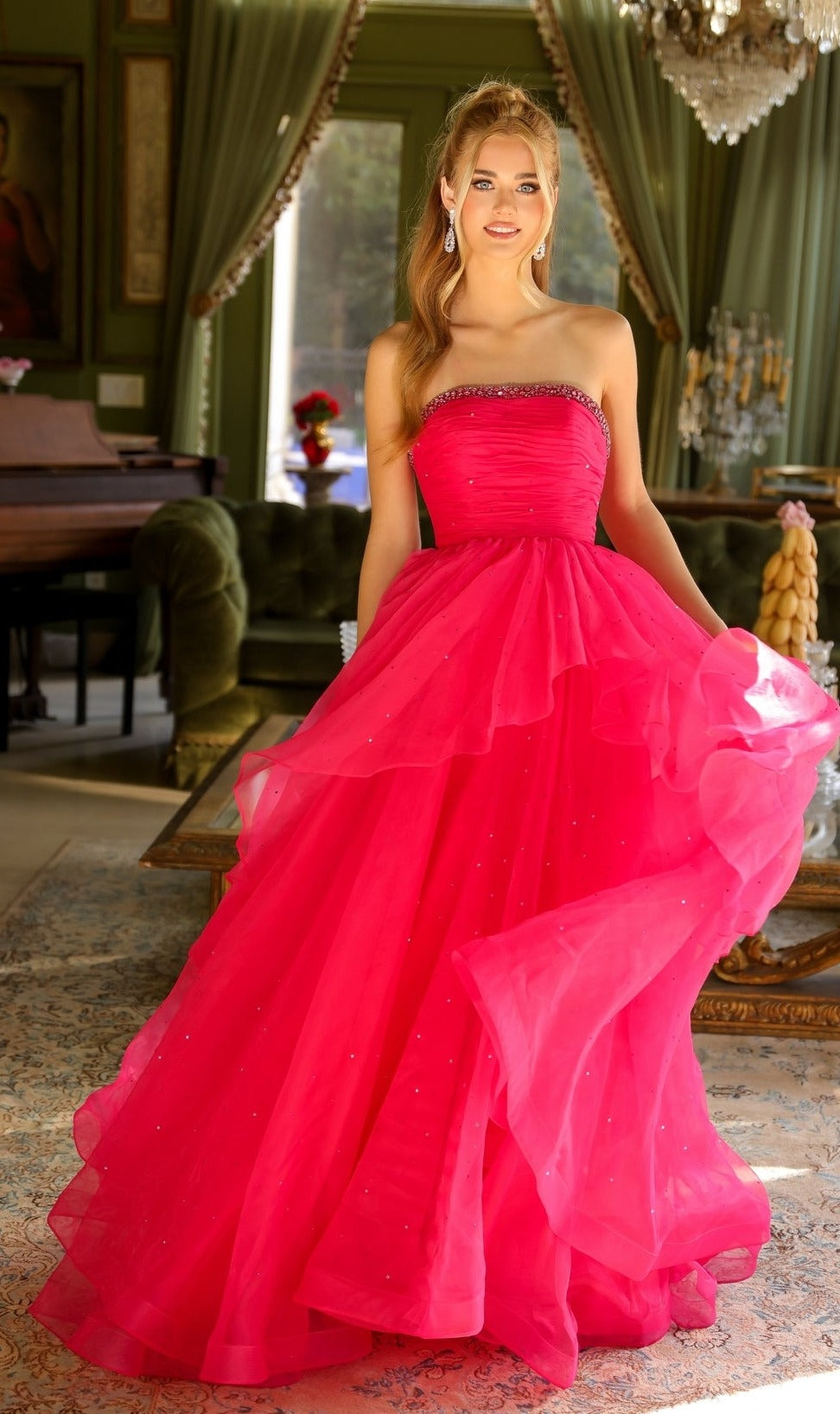 A woman stands indoors in the Ava Presley 29524—a strapless bright pink a-line ball gown with layered tulle and beaded accents—holding the skirt with one hand.