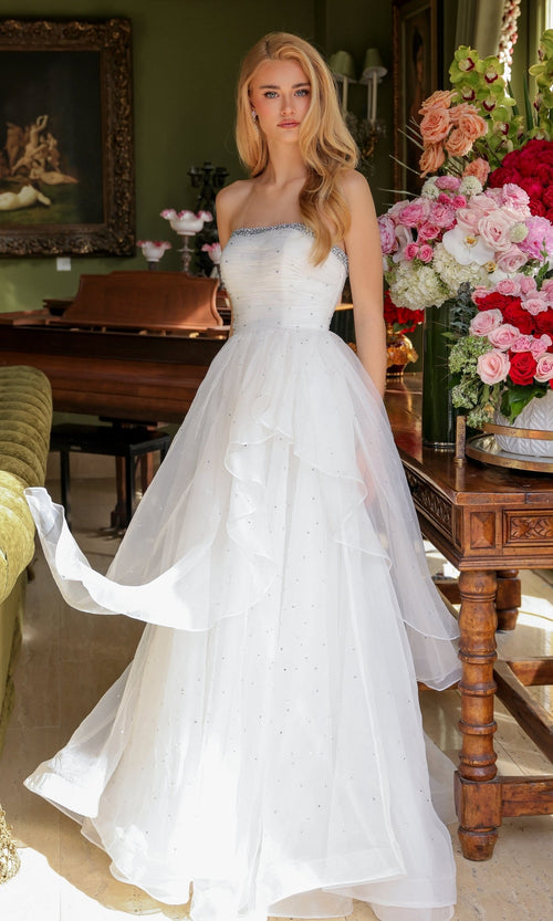 A woman wearing the Ava Presley 29524 Strapless Long Prom Ball Gown stands indoors beside a table of pink and red flowers, with a piano and framed painting in the background.