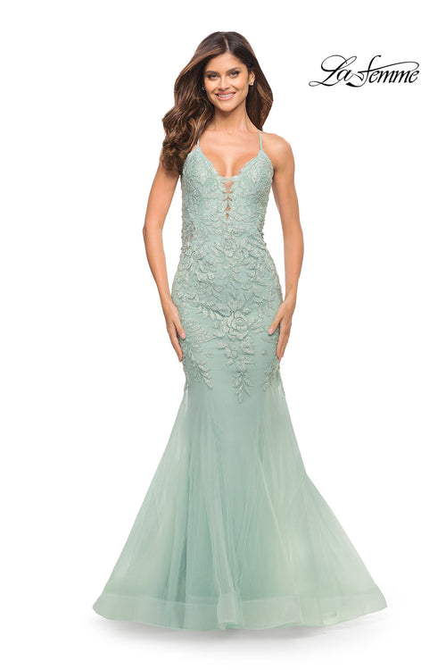 A woman smiles against a plain white background wearing the Backless Long Lace Mermaid Prom Dress by La Femme in light green, featuring floor-length lace, spaghetti straps, and a mermaid silhouette. The "La Femme" logo appears in the top right corner.