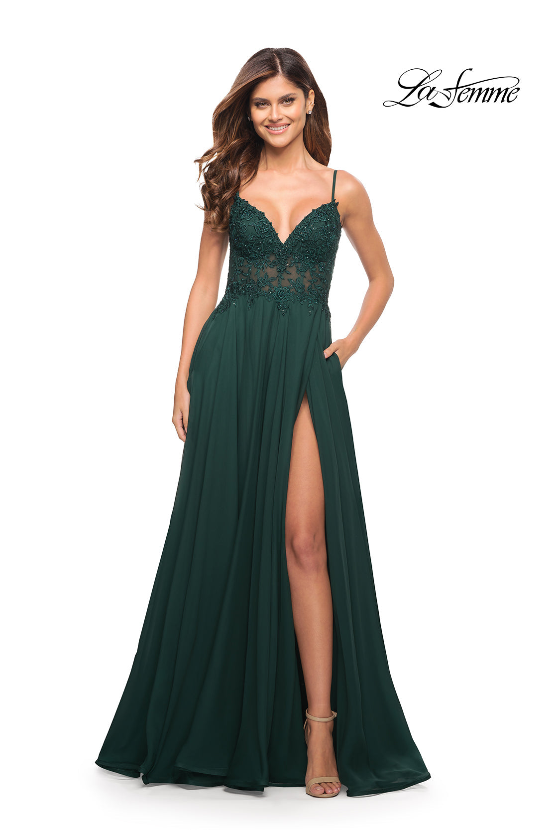 A woman smiles in a dark green La Femme Long A-Line Prom Dress with sheer bodice, thigh-high slit, spaghetti straps, and pockets. The "La Femme" logo is in the top right corner.