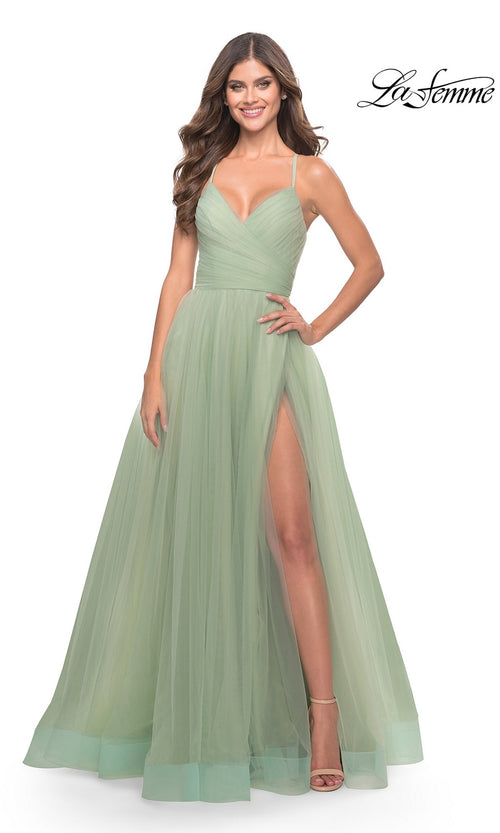 A woman smiles and poses with one hand on her hip, wearing the La Femme Wrap-Bodice Long A-Line Prom Dress 31501 in light green, featuring a fitted bodice, tulle skirt, thigh-high slit, and spaghetti straps.