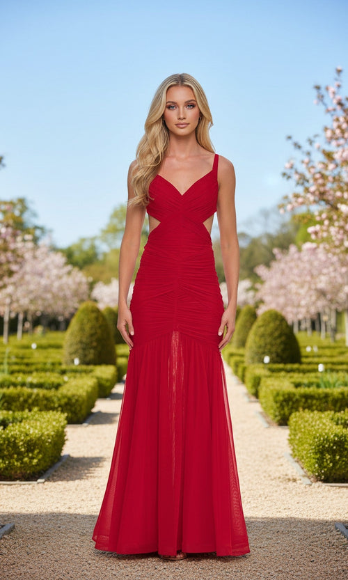 A woman in the La Femme 33626 long red prom dress stands on a gravel path in a manicured garden with blooming trees and trimmed bushes.