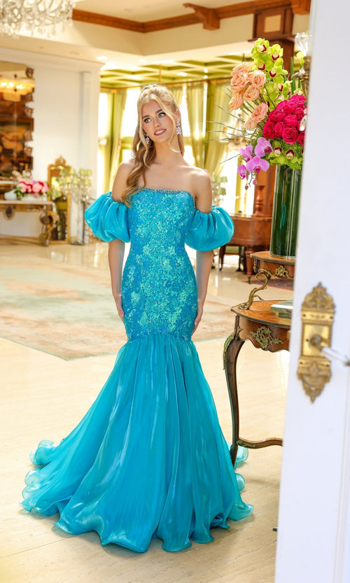 A woman stands indoors by a table with flowers, wearing the Ava Presley 39570—a blue, off-the-shoulder mermaid prom dress featuring puff sleeves.