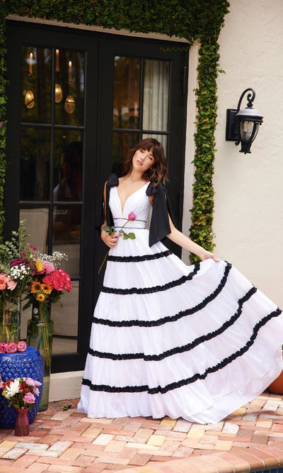 A woman wearing the Ava Presley 40091 Shoulder Bows A-Line Formal Dress poses outdoors by glass doors, holding a pink rose. Floral vases are arranged nearby on the brick patio.