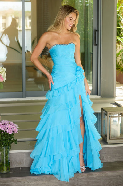 A woman in the strapless Ava Presley 42242 ruffled prom gown in sky blue stands on steps by a glass door, holding the side of her evening dress and smiling.