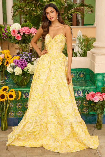 A woman wears the Ava Presley 42312 long yellow prom dress with an elegant A-line skirt, standing outdoors among green tiles, columns, and vibrant flowers.