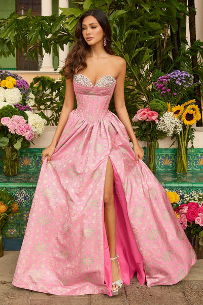 A woman in the Ava Presley 42318 long prom dress, hot pink with a strapless corset bodice and thigh-high slit, stands before vibrant flowers and greenery.