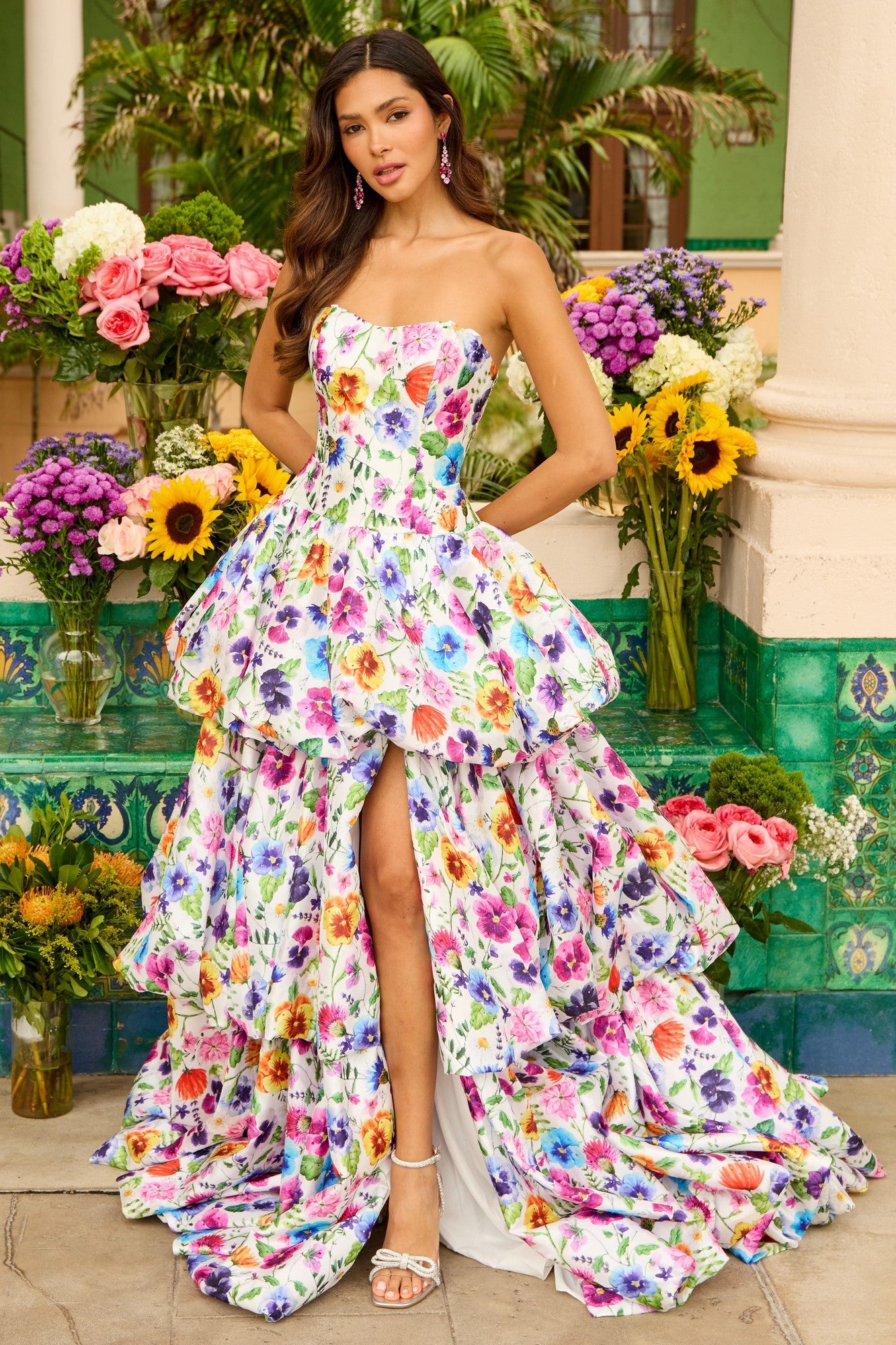 A woman stands among flowers wearing the Ava Presley 42360 strapless print prom ball gown in White/Multi, featuring a long silhouette with a high slit and vibrant floral design.