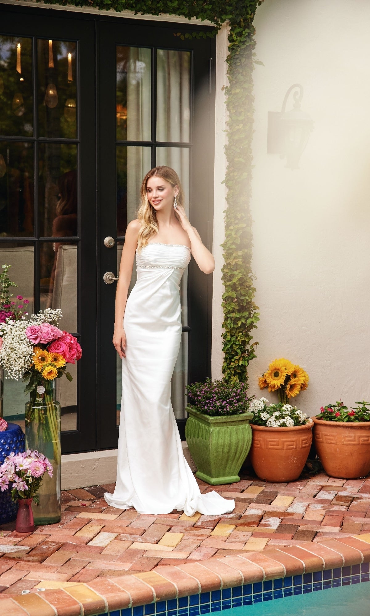 A woman in the Ava Presley 47026 long prom dress stands by a glass door, surrounded by potted plants and flowers beside a pool on a brick patio.