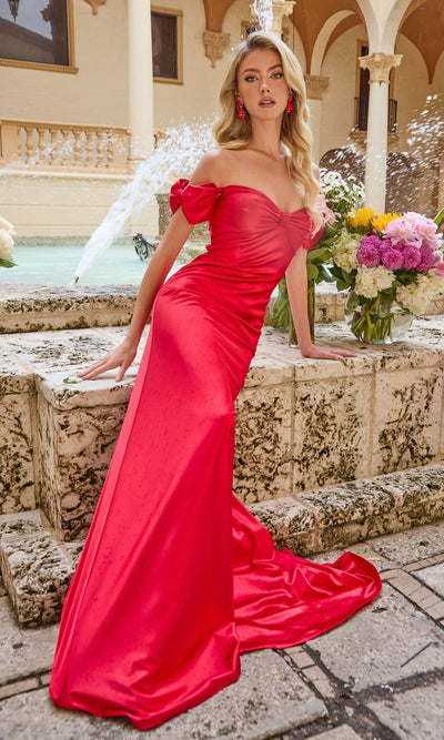 A woman in a red Off-Shoulder Satin Prom Gown (Ava Presley 47178) sits on a stone ledge near a fountain, surrounded by lush floral arrangements in an elegant outdoor courtyard.