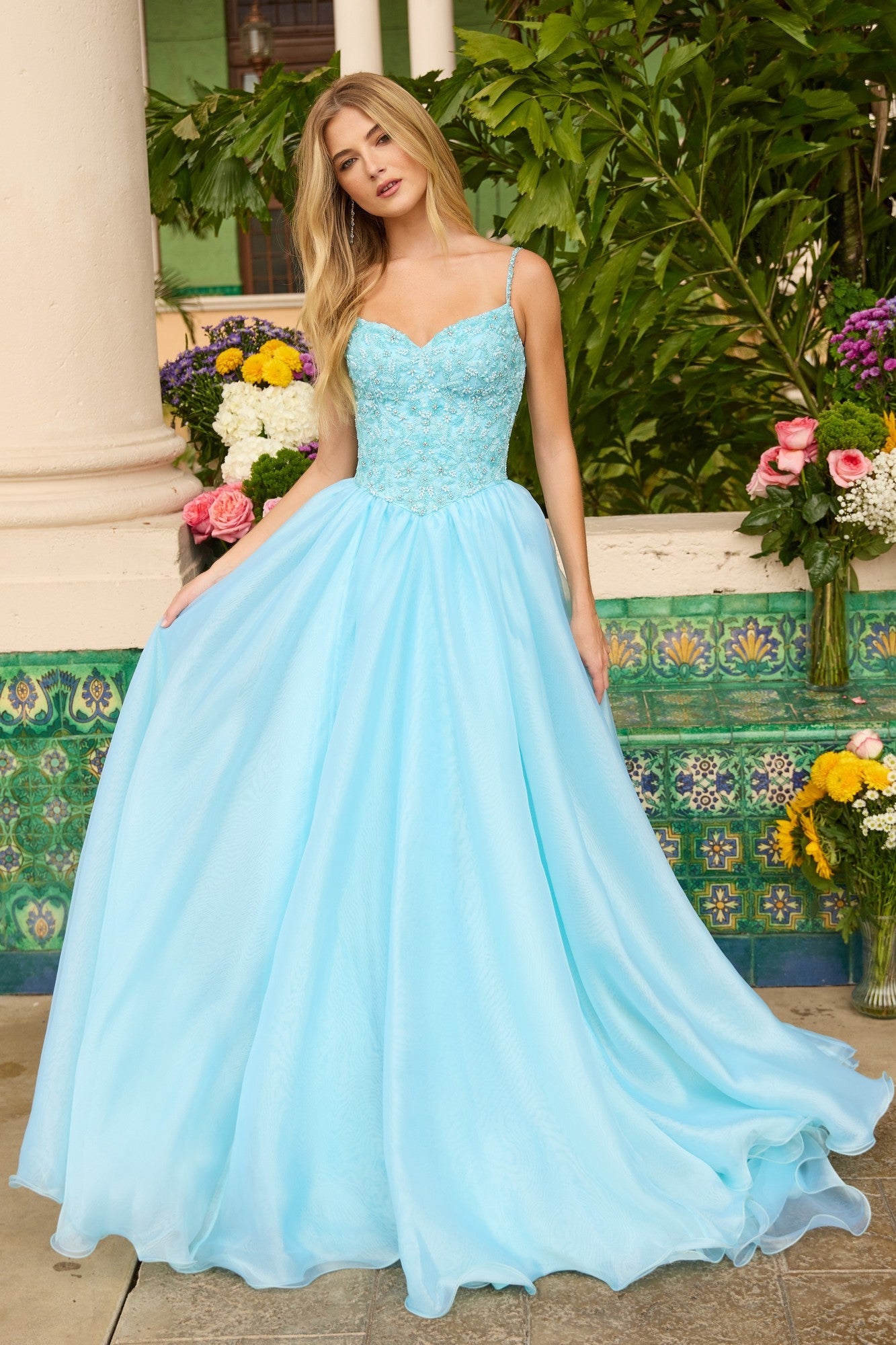 A woman stands outdoors near flowers and greenery wearing the Ava Presley 47242 long prom dress in light blue, featuring an a-line skirt and thin straps.