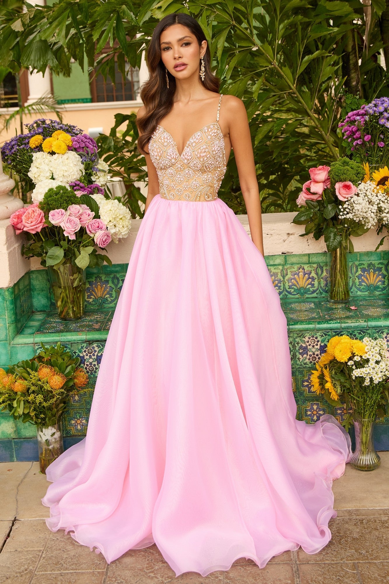 A woman in the Ava Presley 47243 long prom dress in nude/light pink, featuring a beaded bodice, stands outdoors among colorful flowers and lush green plants.