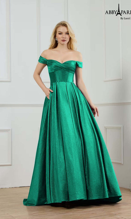 A woman stands in a white paneled room wearing the Abby Paris 90373, a long emerald prom dress featuring off-the-shoulder sleeves, shimmer satin fabric, and a corset bodice.