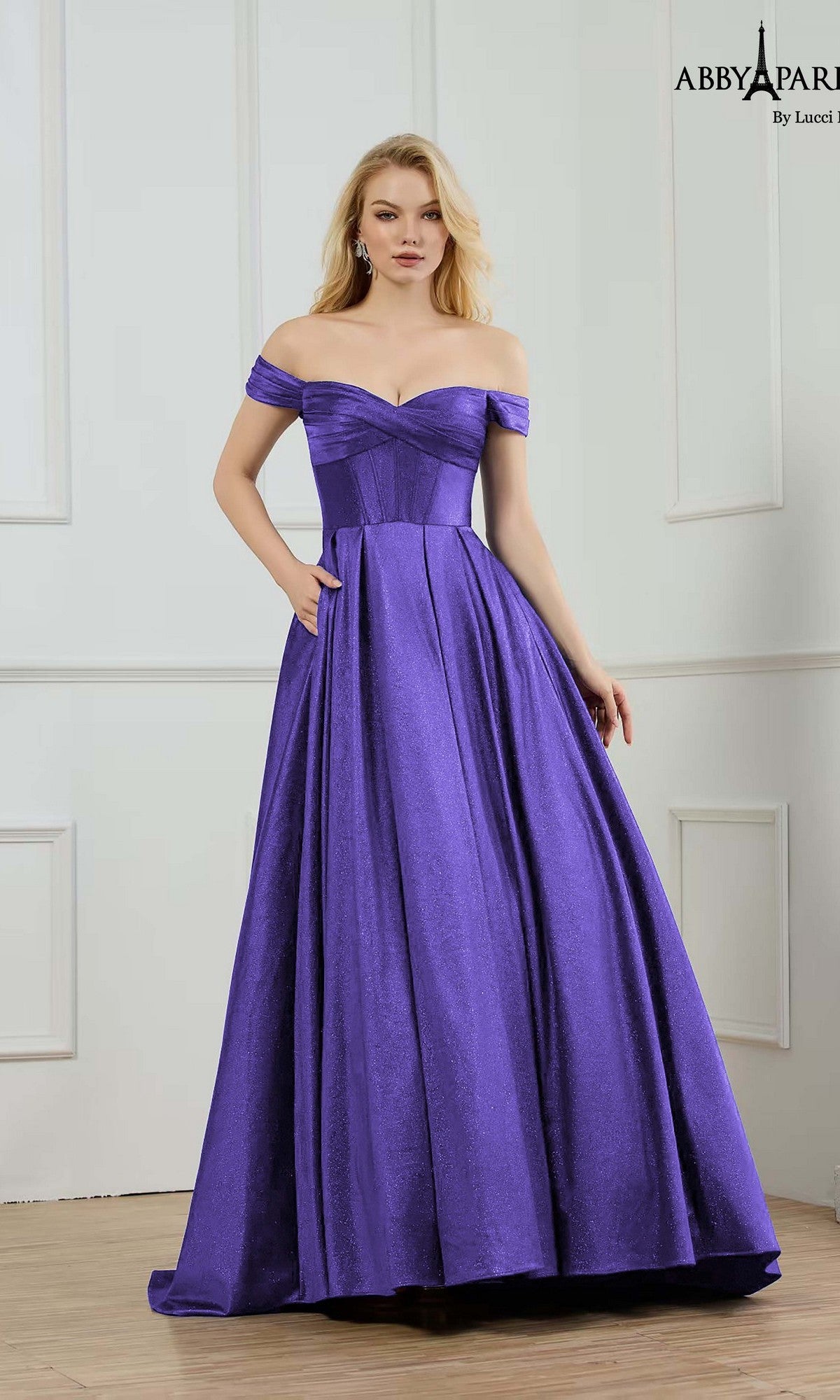 A woman stands in a light-colored room with paneled walls, wearing the Abby Paris 90373 long purple prom dress featuring an off-the-shoulder corset bodice and floor-length shimmer satin.
