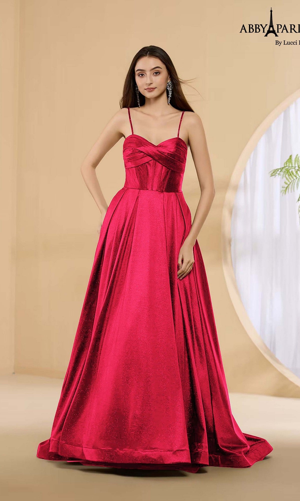 A woman in the Abby Paris 90389 long red prom dress with spaghetti straps and a sleeveless, floor-length satin design stands in a minimalist room with beige walls and a round window.