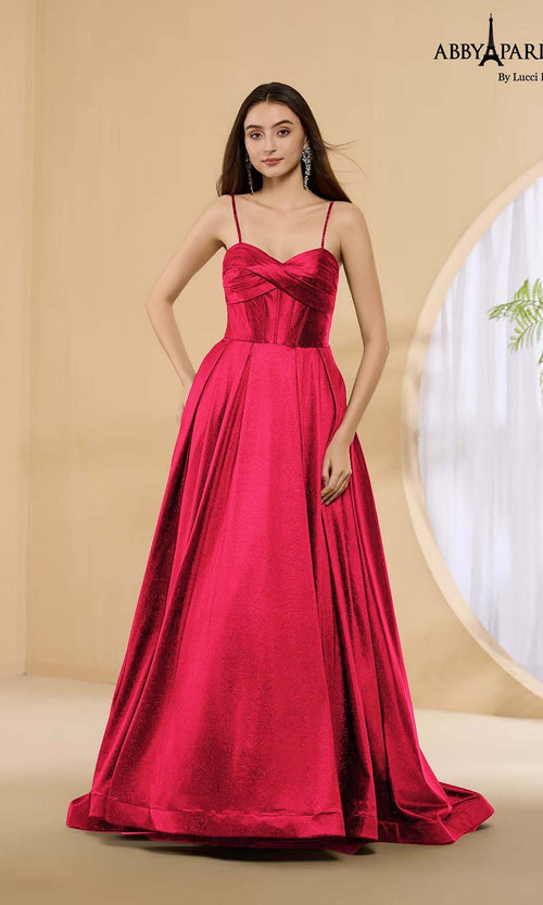 A woman in the Abby Paris 90389 long red prom dress with spaghetti straps and a sleeveless, floor-length satin design stands in a minimalist room with beige walls and a round window.