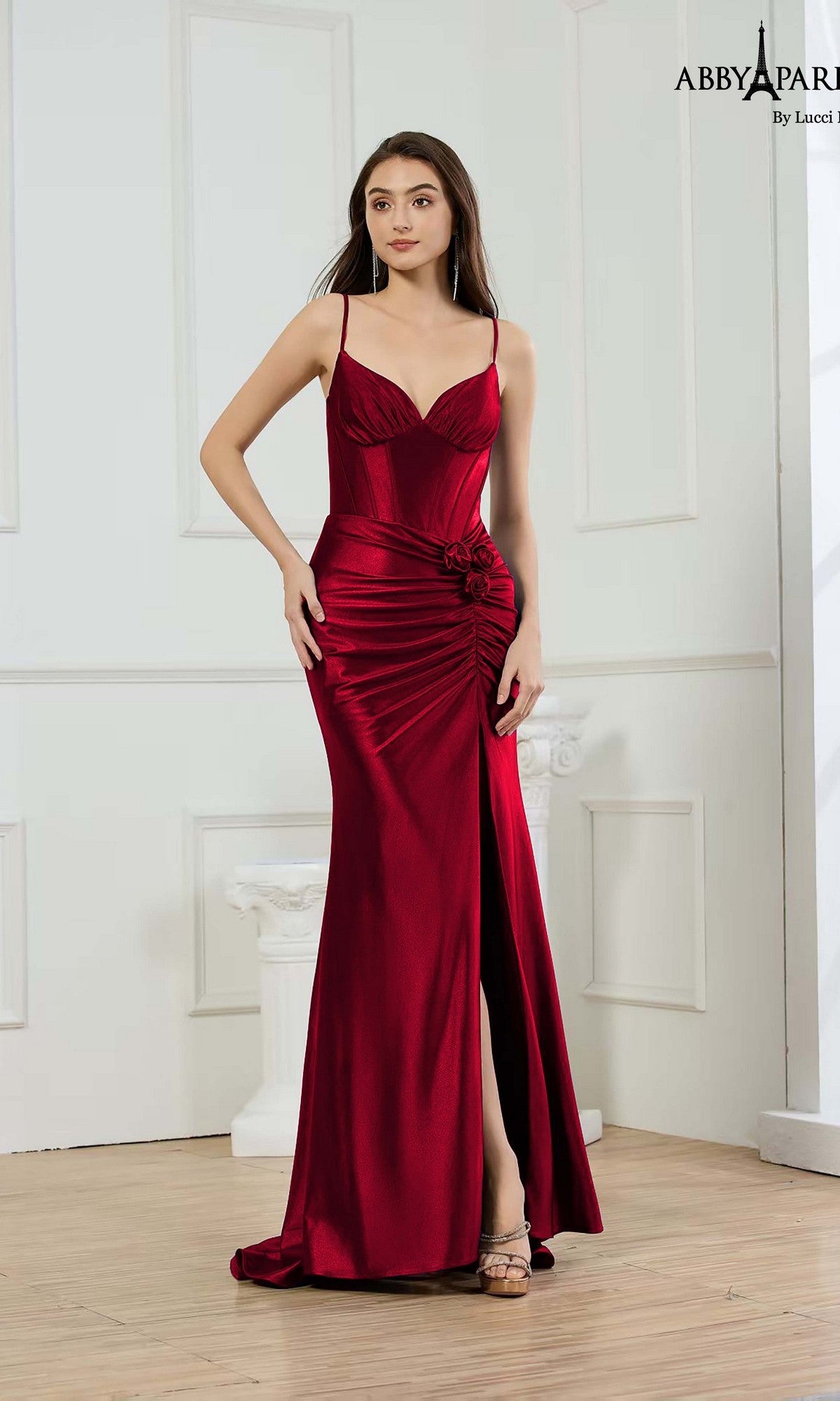 A woman stands indoors wearing the Abby Paris 90393 long prom dress in wine, featuring shiny stretch satin, spaghetti straps, a ruched waist, and a thigh-high slit.