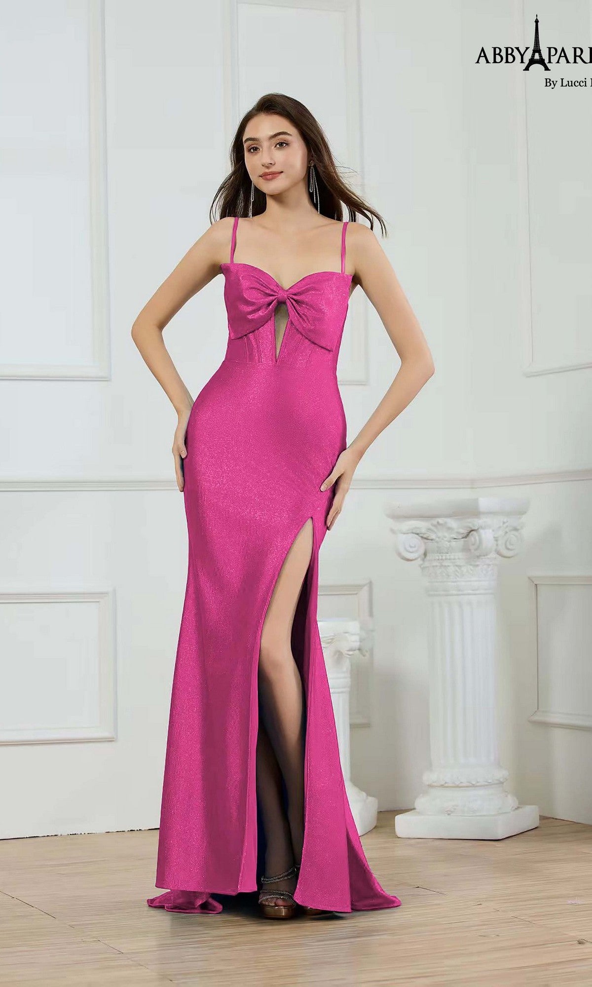 A woman stands indoors beside a white column, wearing the fuchsia Abby Paris 90396 long prom dress featuring stretch satin, a corset bodice, front bow detail, and a high-leg side slit.