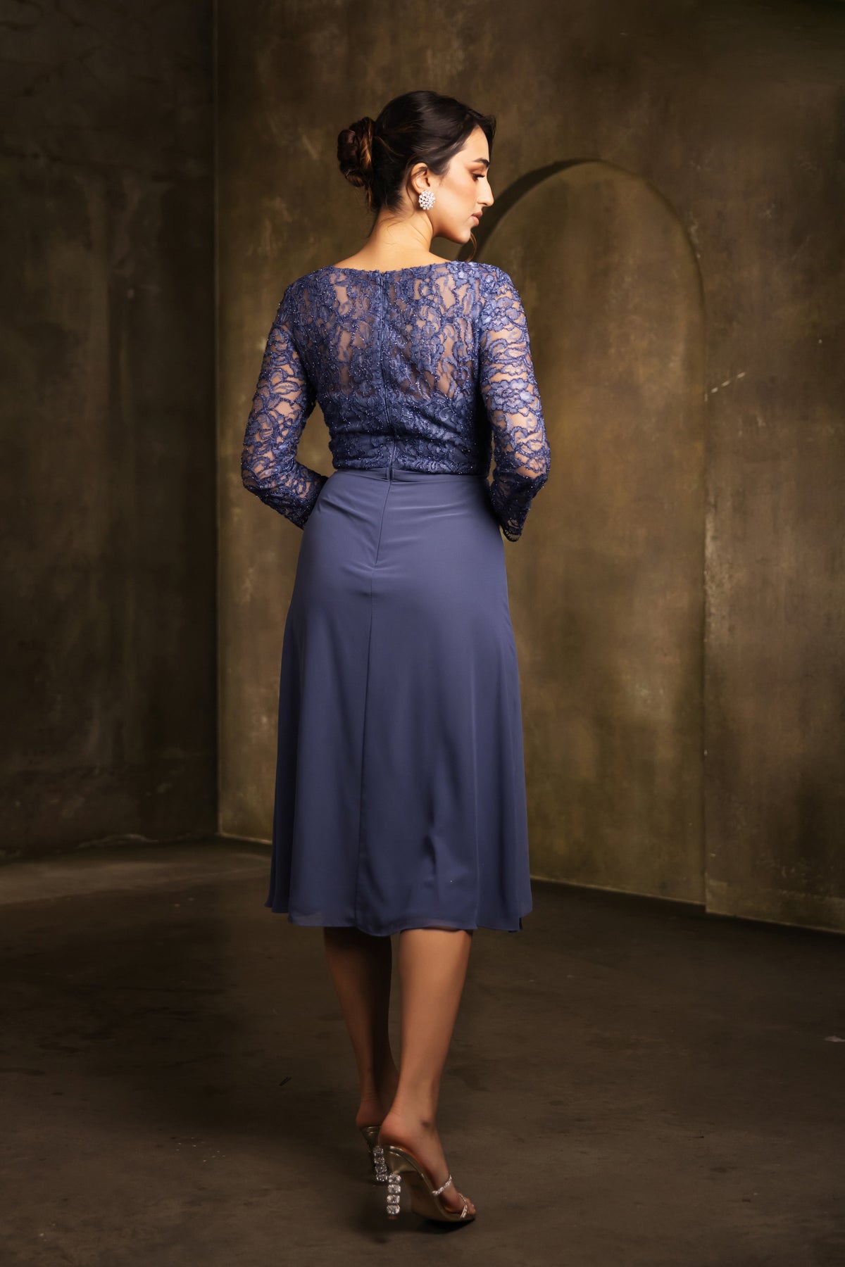 A woman wearing a navy blue lace long-sleeve midi dress, shown from the back in a studio setting with a textured dark brown wall background.
